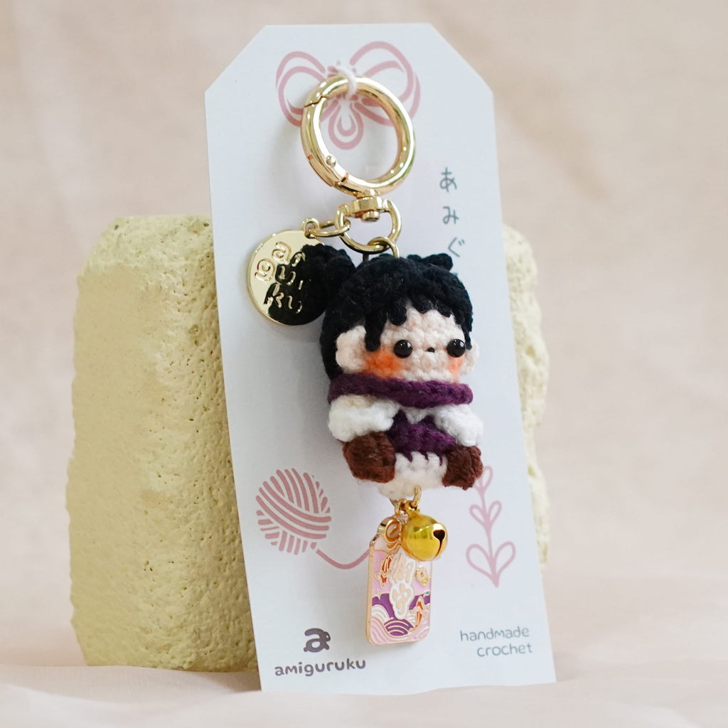 A small plush keychain with a cute character hanging from a gold ring, attached to a white branded card and displayed on a textured beige fabric surface.