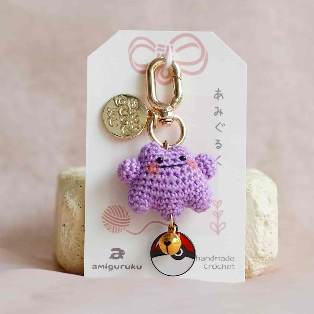 A small crocheted purple monster keychain charm with gold-tone hardware, displayed on a white branded card insert against a soft pink fabric background.
