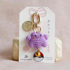 A small crocheted purple monster keychain charm with gold-tone hardware, displayed on a white branded card insert against a soft pink fabric background.