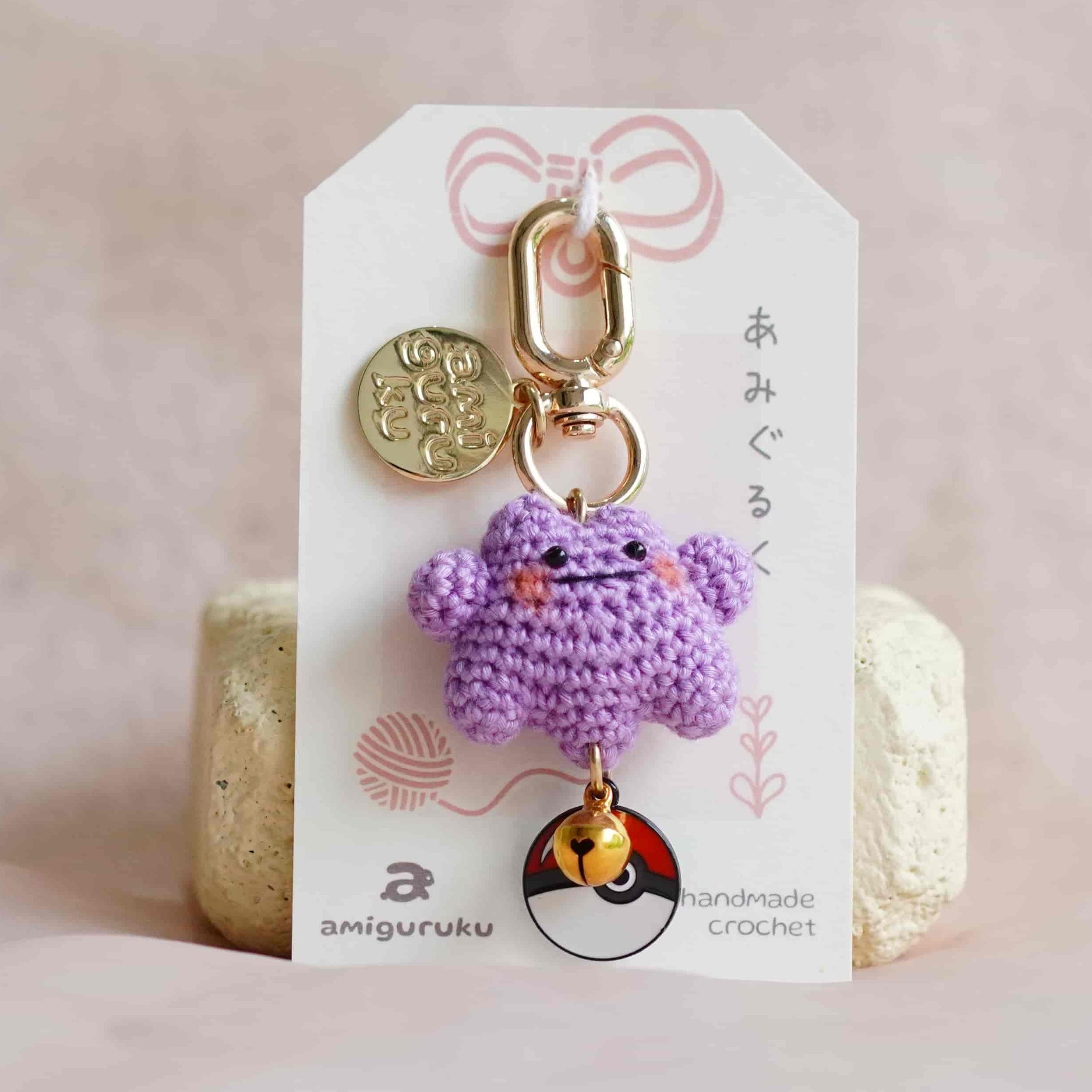 A small crocheted purple monster keychain charm with gold-tone hardware, displayed on a white branded card insert against a soft pink fabric background.
