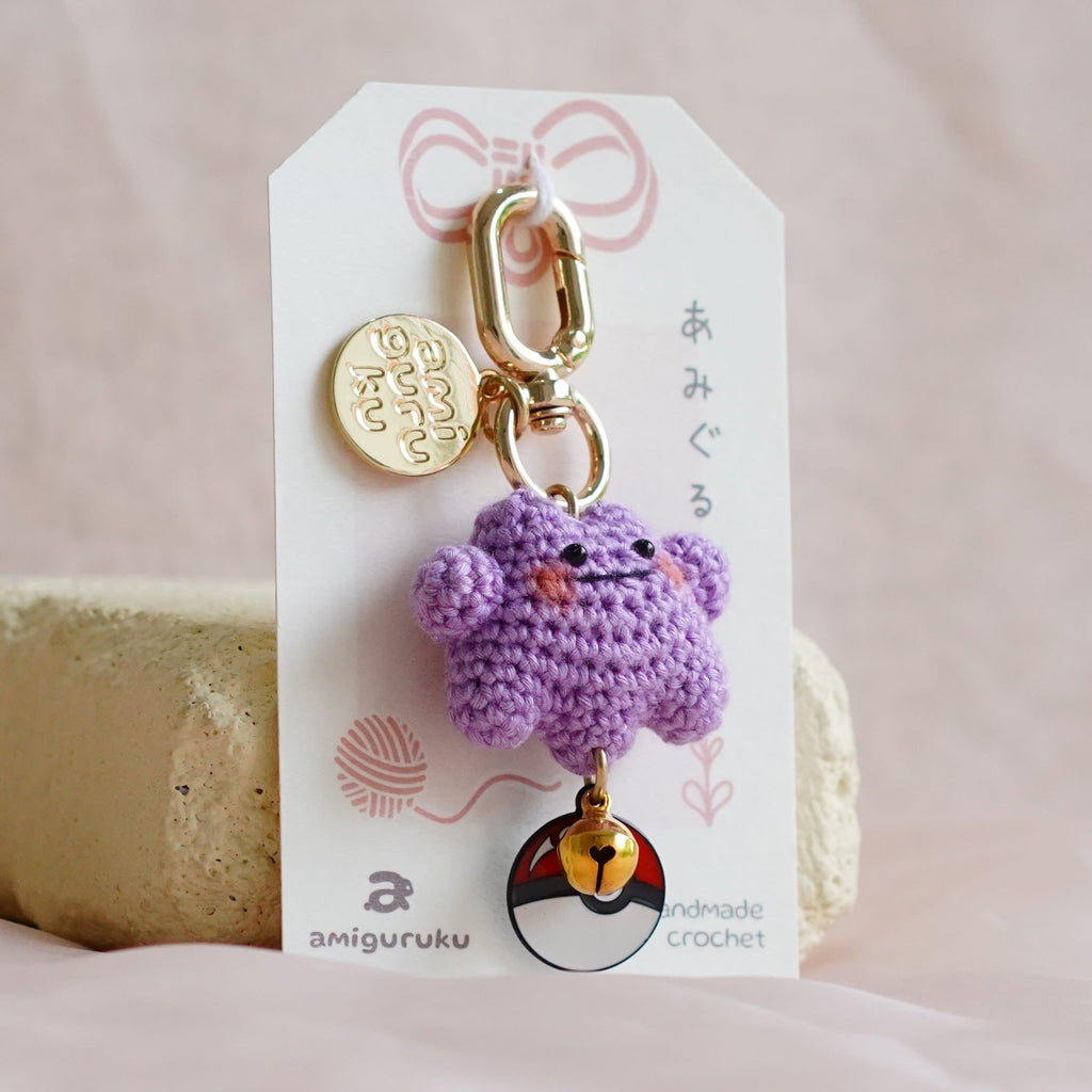 A small purple crocheted character charm keychain with gold-tone hardware, attached to a white display card, resting on a beige block against a soft pink fabric backdrop.