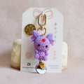 A small handmade crocheted purple character charm with a gold clasp and a Pokéball bell, attached to a white display card, photographed on a soft pink fabric background.