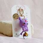 A small purple crocheted keychain charm with gold-tone hardware and a tiny multicolor accent, attached to a white display card and photographed on soft beige fabric.