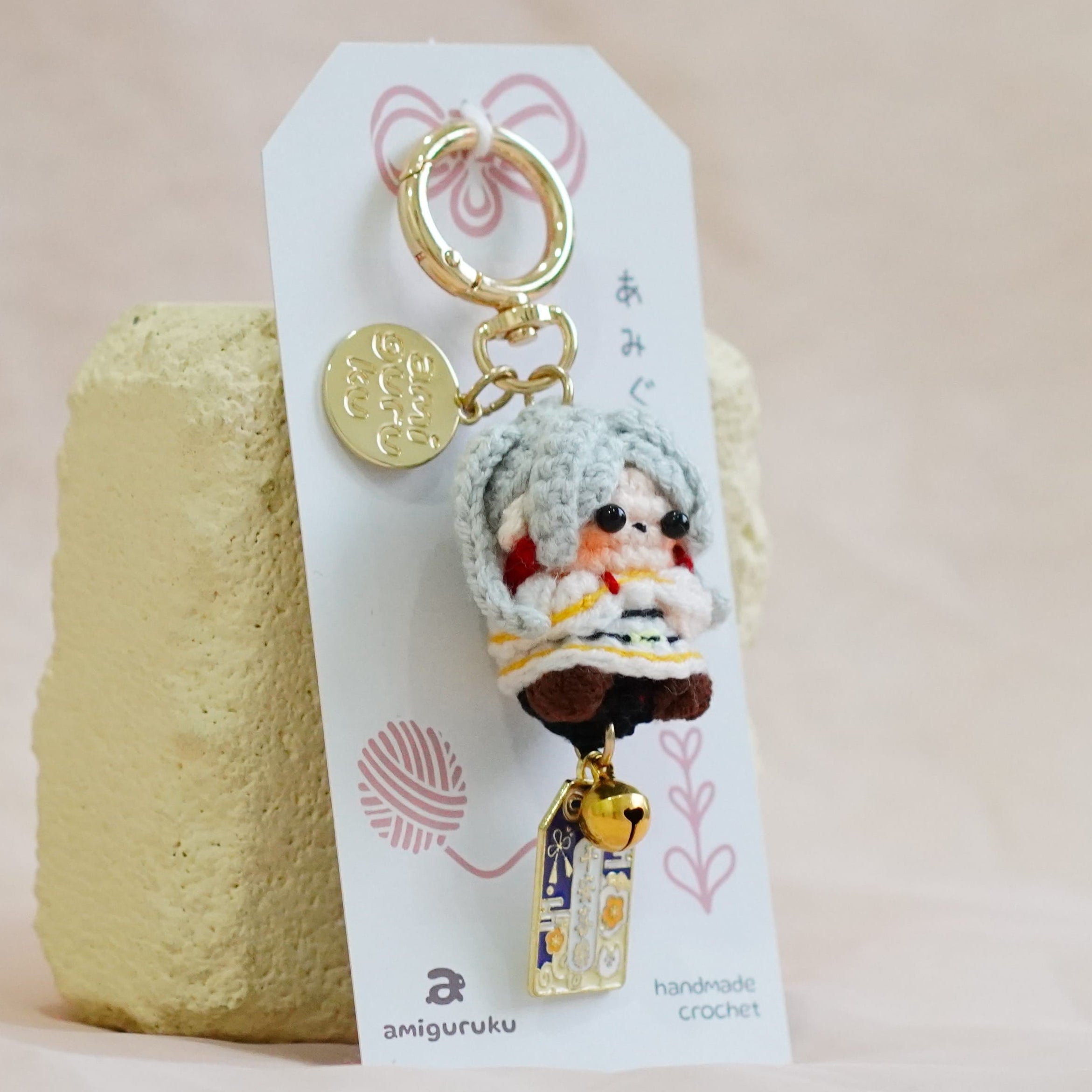 A small character keychain with a white plush figure and gold-tone hardware, attached to a printed display card and standing on a textured beige surface.