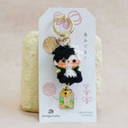 A small anime keychain charm featuring a black-haired character in a black and white outfit with a gold bell, attached to a white display card and standing in front of a textured beige background.