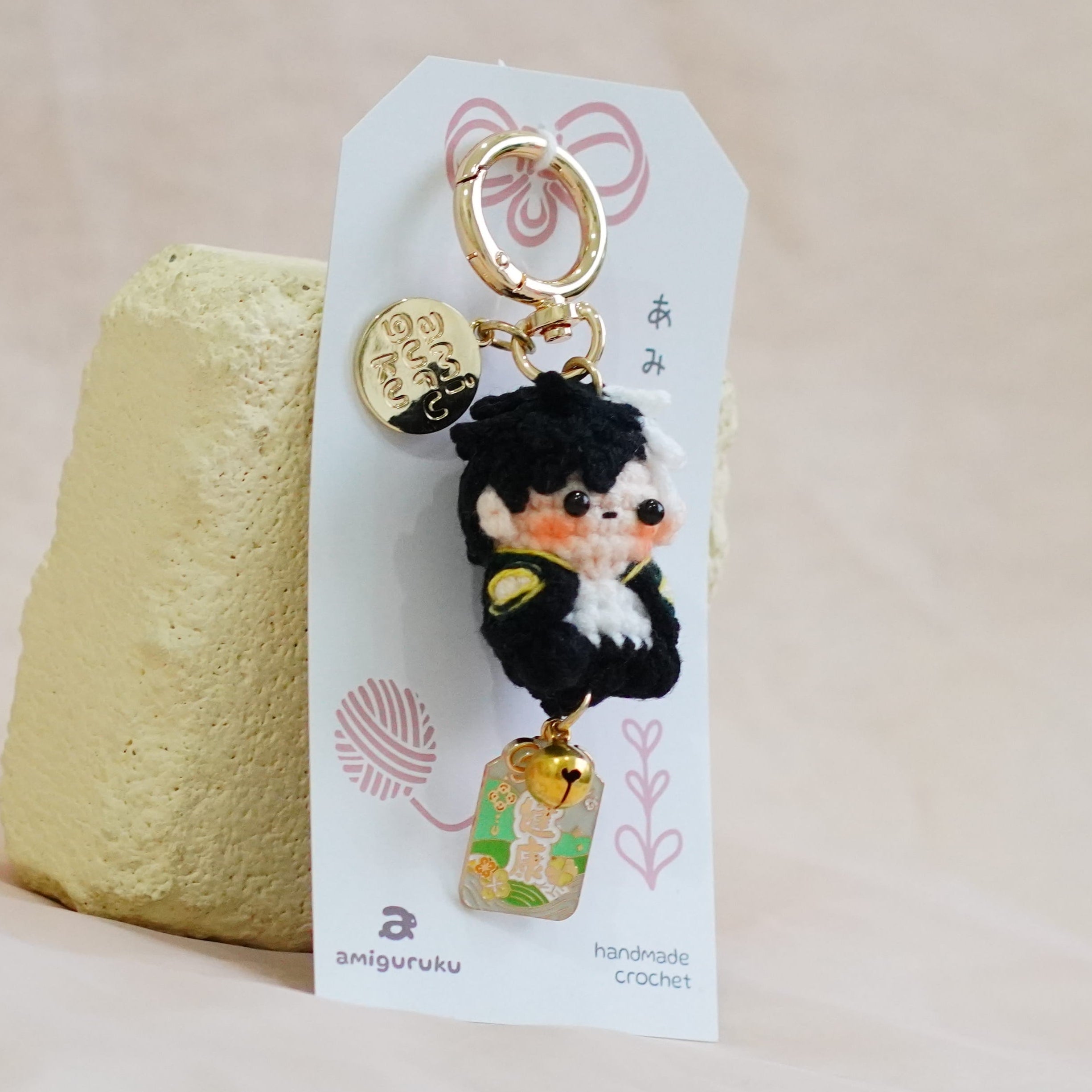 A small black-and-white plush character keychain with gold-tone hardware, attached to a white decorative card, displayed on a soft beige fabric surface next to a pale yellow textured object.