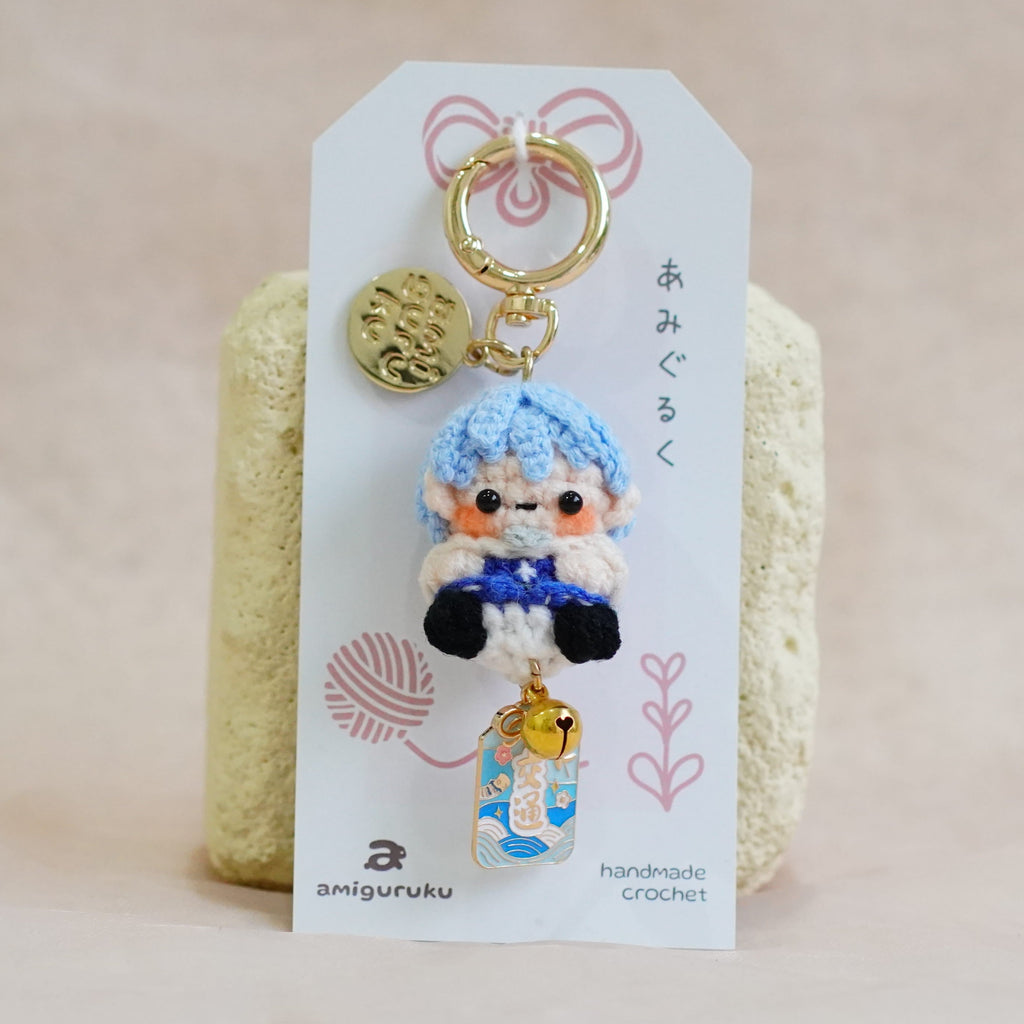 A handmade crocheted keychain featuring a small blue-haired character doll, attached to a white display card with Japanese text and a gold key ring, set against a soft beige fabric background.