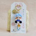 A handmade crocheted keychain featuring a small blue-haired character doll, attached to a white display card with Japanese text and a gold key ring, set against a soft beige fabric background.