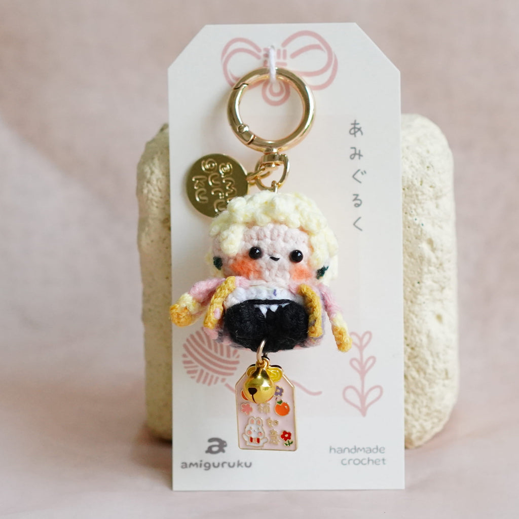 A handmade crocheted doll keychain with blonde hair, a black dress, and gold accessories, attached to a white display card with Japanese text, set against a soft pink fabric background.