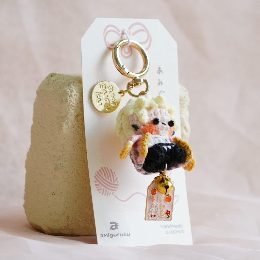 A small plush keychain or bag charm with a cute character and gold key ring, attached to a white display card and propped against a textured beige stone on soft pink fabric.