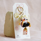 A small plush keychain or bag charm with a cute character and gold key ring, attached to a white display card and propped against a textured beige stone on soft pink fabric.