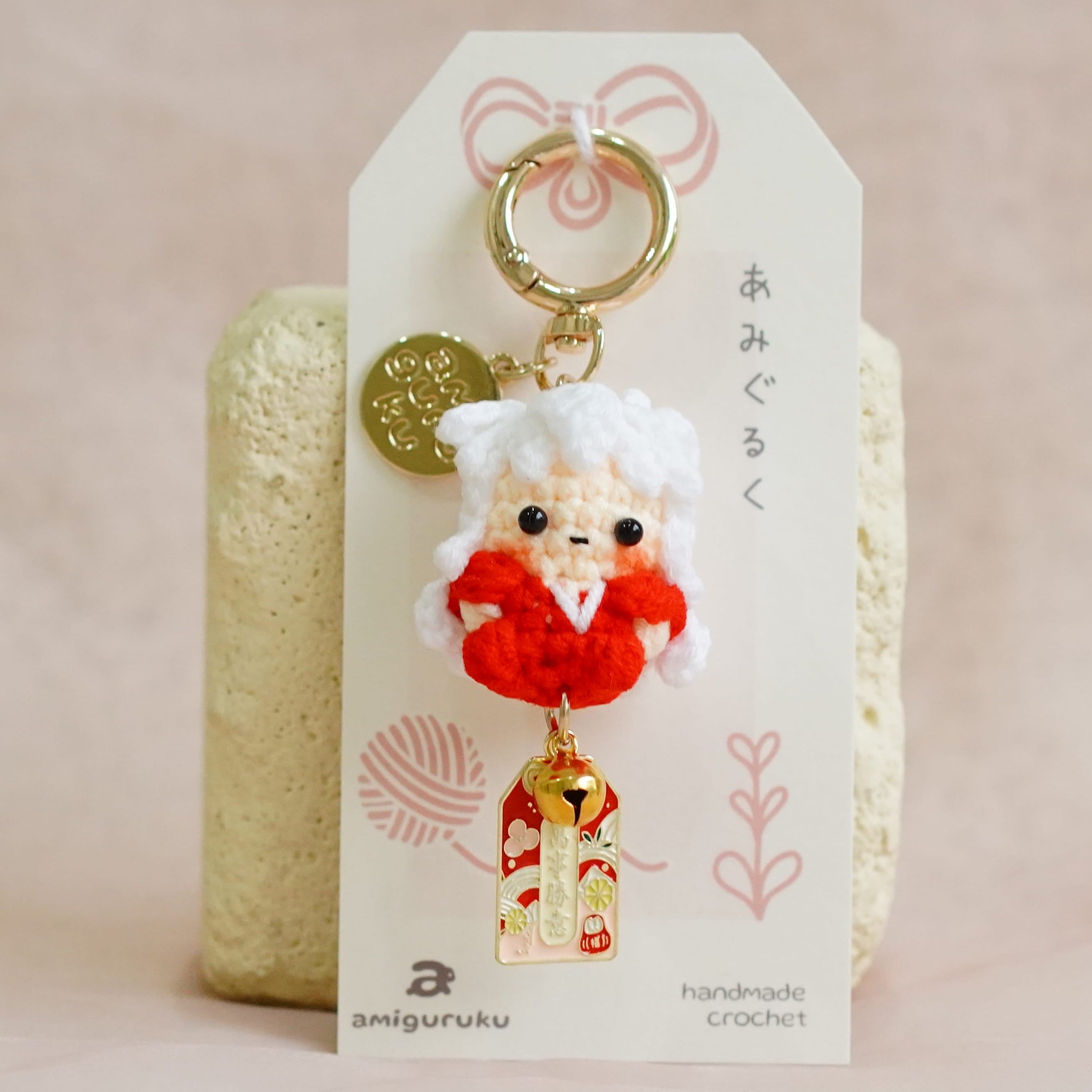 A handmade Japanese Amigurumi keychain featuring a small red and white plush Santa Claus figure with a gold keyring and charm, displayed on a branded white card over a gold textured stand against a soft beige background.
