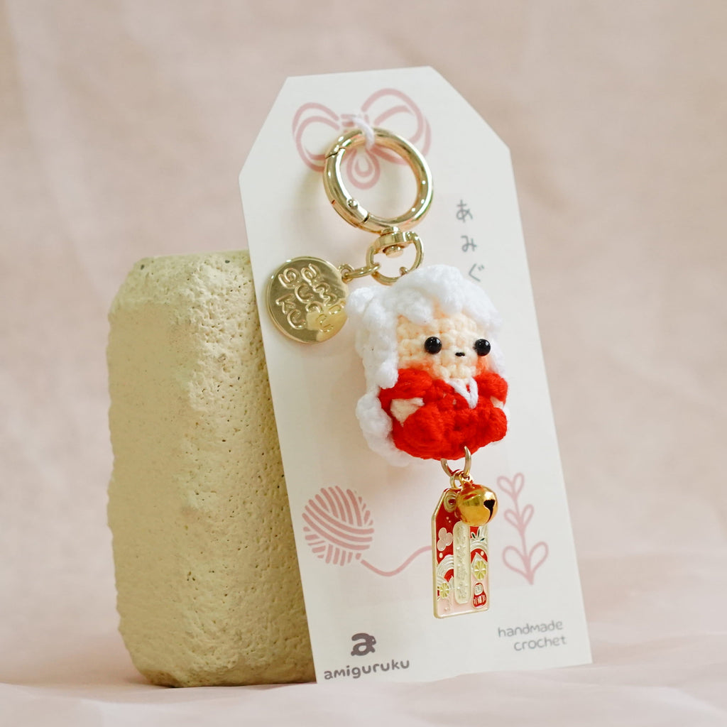 A small hanging keychain or bag charm with a white fluffy character wearing a red outfit, attached to a white card and displayed next to a textured beige stone on soft cream fabric.