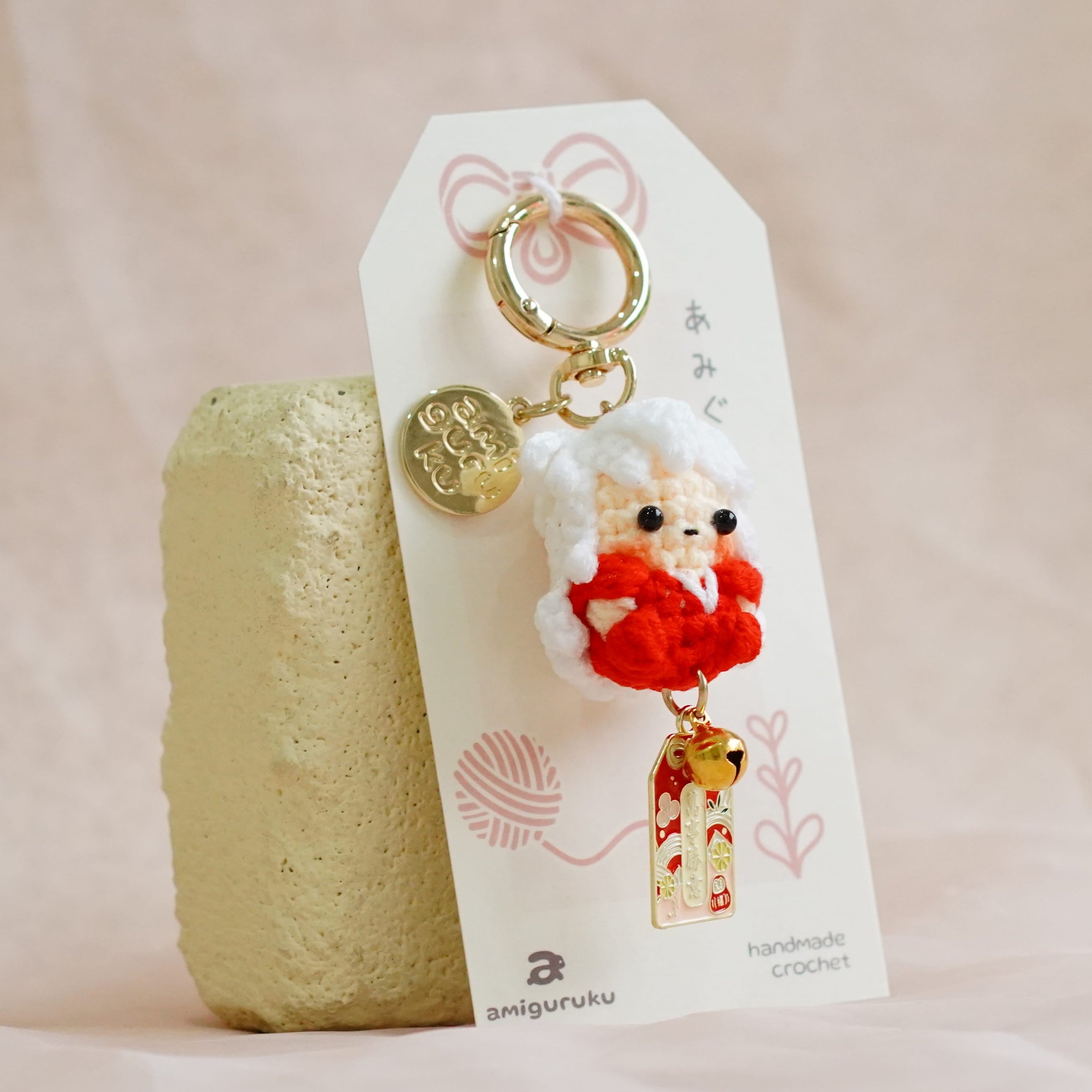A small hanging keychain or bag charm with a white fluffy character wearing a red outfit, attached to a white card and displayed next to a textured beige stone on soft cream fabric.