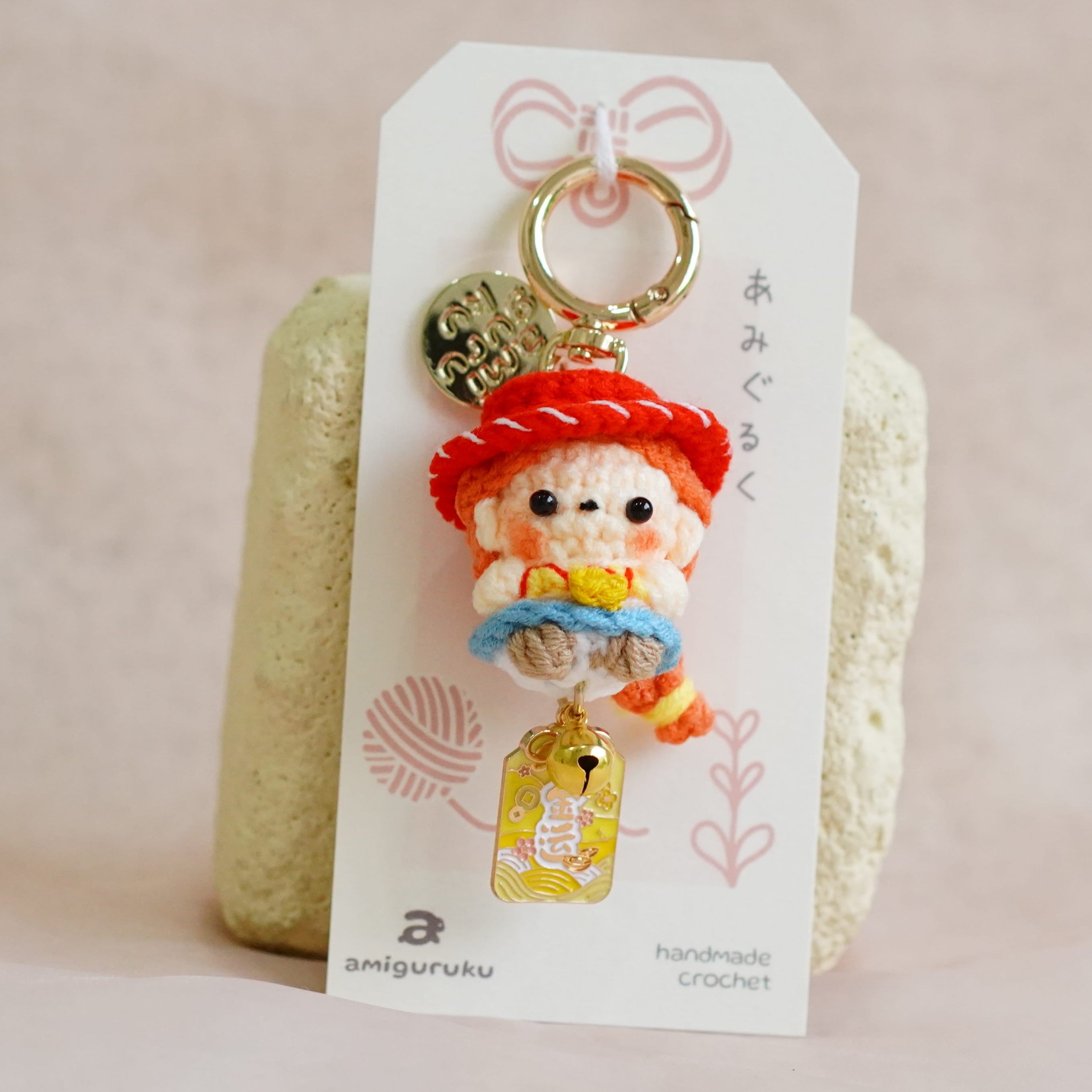 A small handmade charm featuring a cartoon character with a red hat and blue outfit, attached to a white display card with a gold key ring, photographed against a soft beige fabric background.