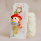 A small plush keychain featuring a character in a red hat and blue dress, attached to a white display card with a gold keyring, photographed against a soft beige fabric backdrop.