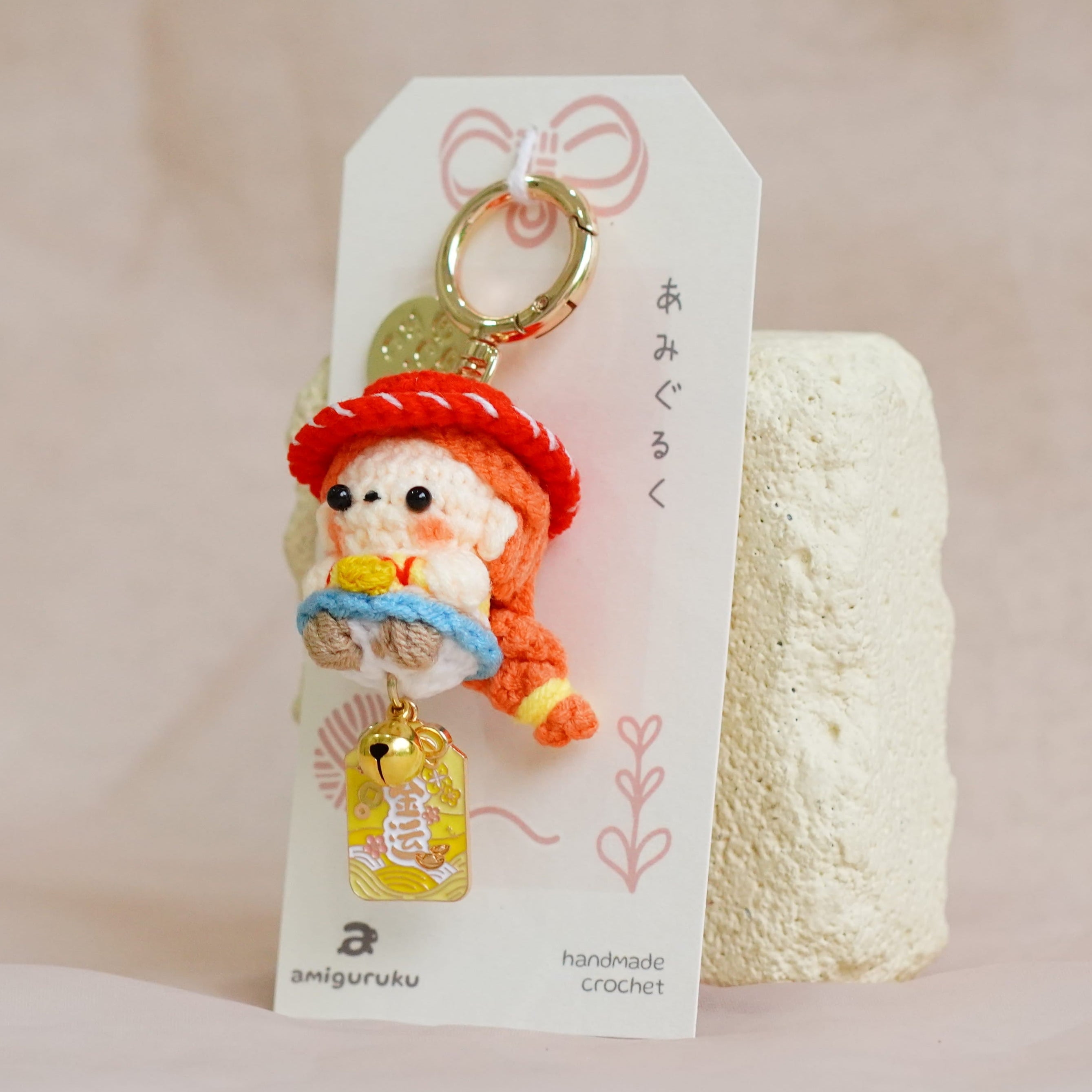 A small plush keychain featuring a character in a red hat and blue dress, attached to a white display card with a gold keyring, photographed against a soft beige fabric backdrop.