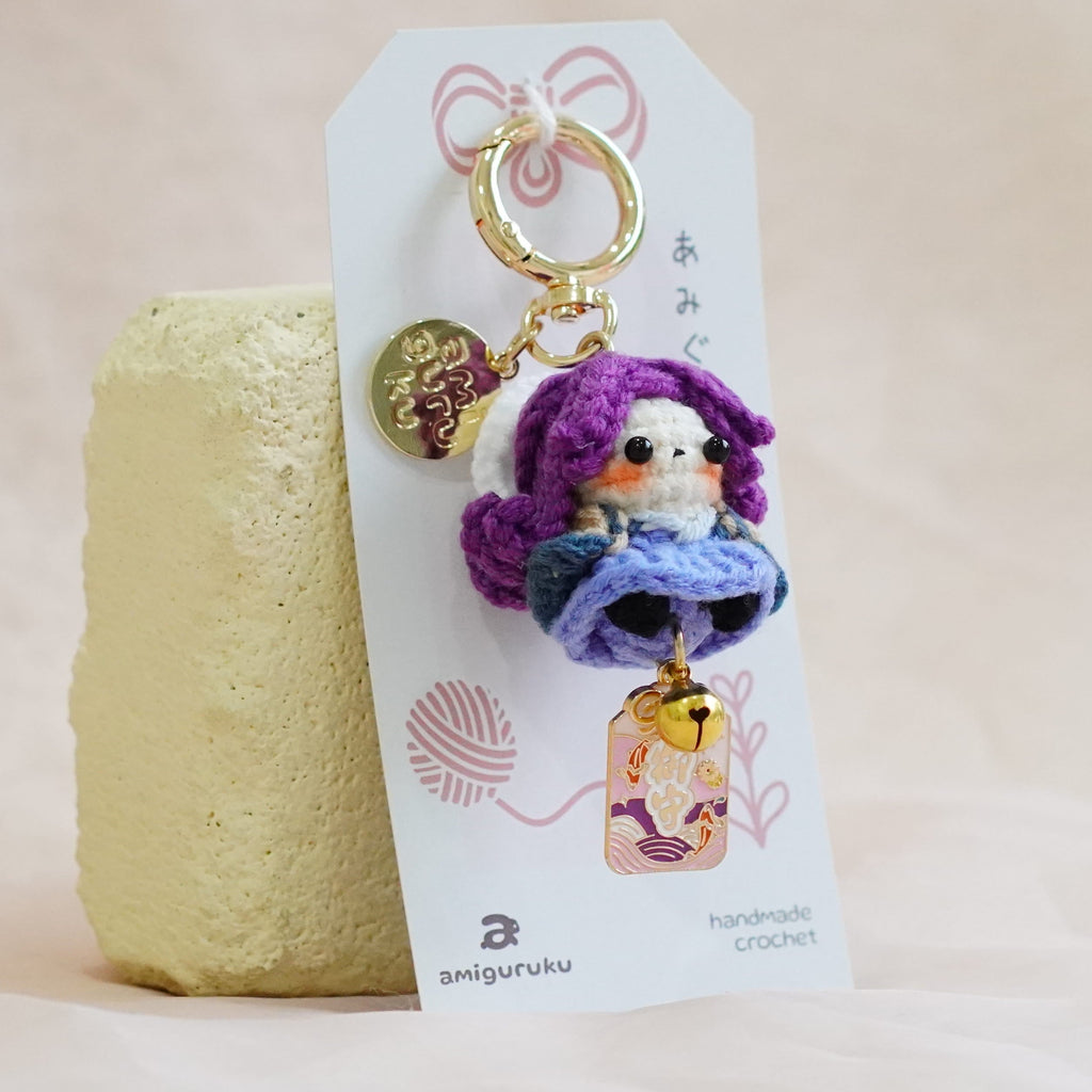 A handmade crochet keychain featuring a small bird character with purple hair and a blue dress, attached to a white display card and standing next to a textured yellow candle on a soft beige fabric background.