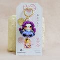 A handmade crocheted keychain charm featuring a purple-haired doll in a blue dress with a small gold bell, attached to a white display card with Japanese text and a pink bow, set against a soft beige fabric background.