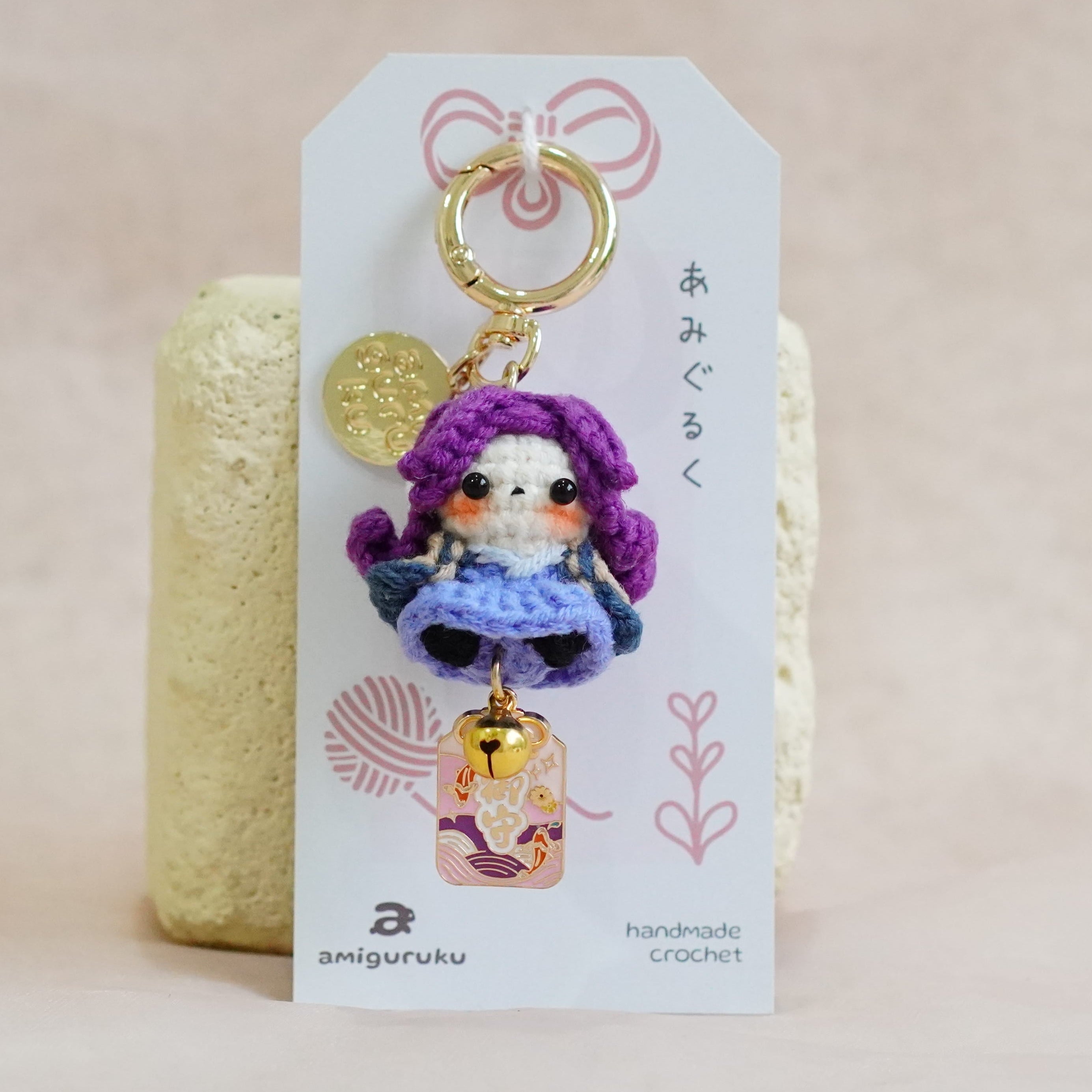 A handmade crocheted keychain charm featuring a purple-haired doll in a blue dress with a small gold bell, attached to a white display card with Japanese text and a pink bow, set against a soft beige fabric background.