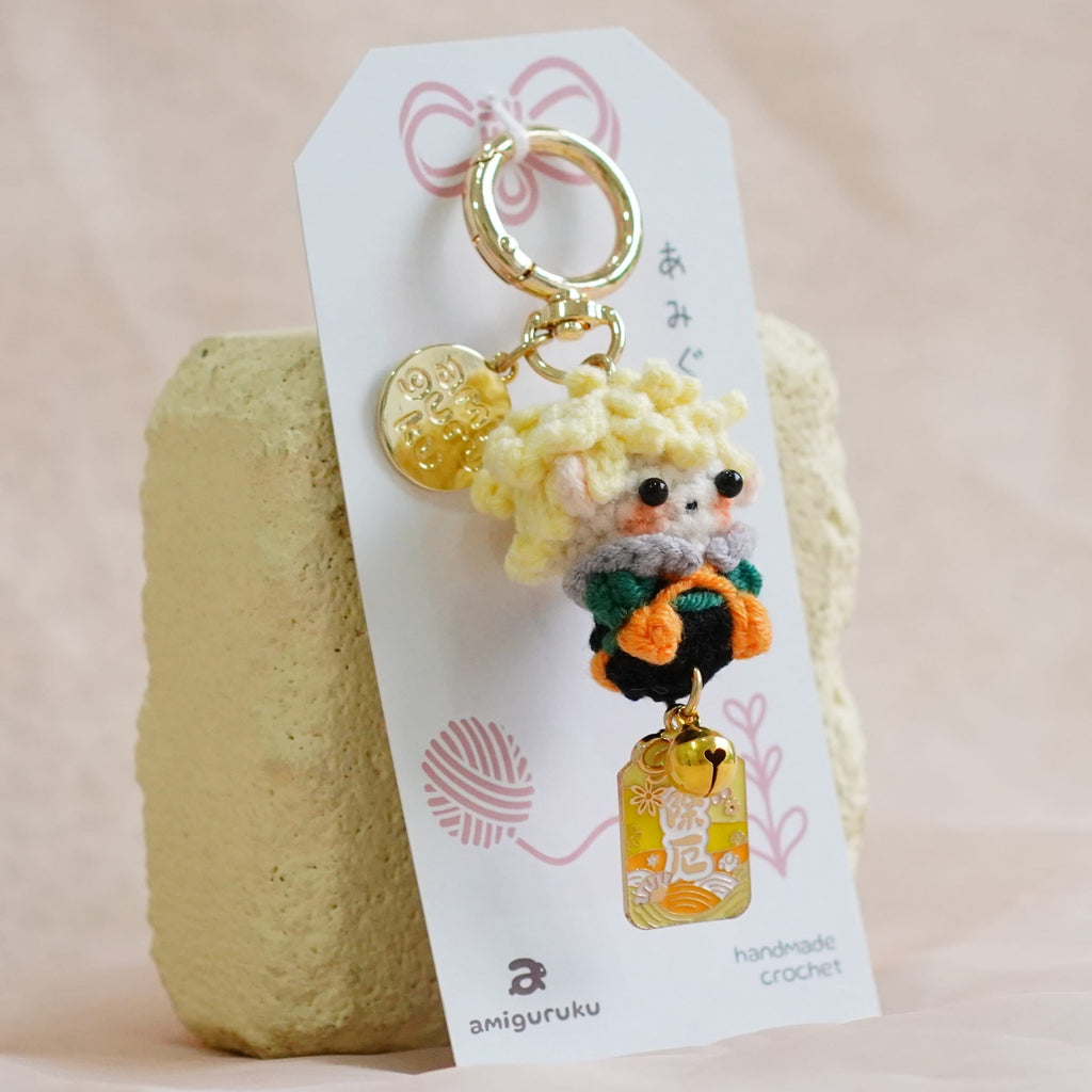 A handmade crochet keychain featuring a small dog character in an orange outfit with a yellow flower, attached to a white paper tag and displayed on a textured beige fabric surface.