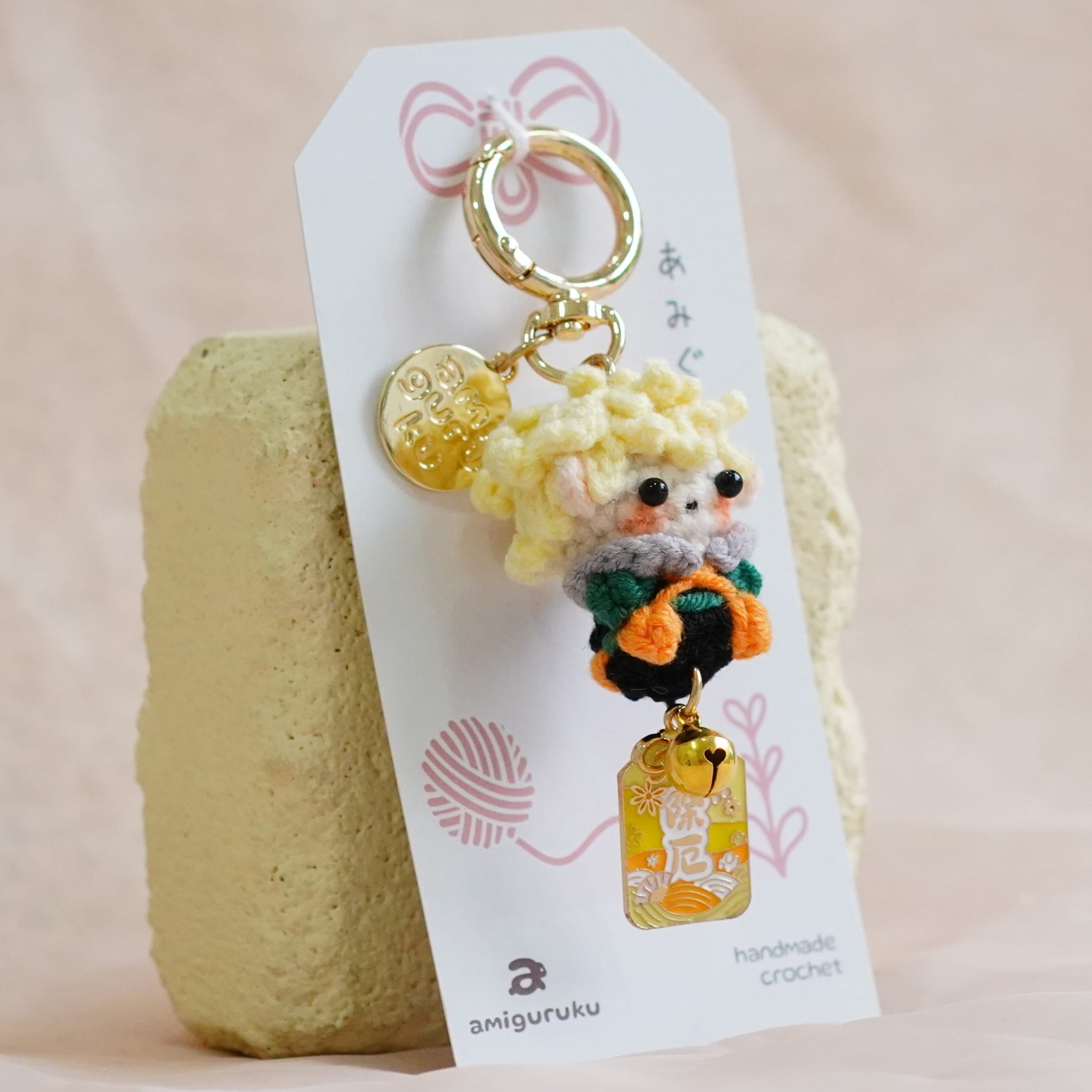A handmade crochet keychain featuring a small dog character in an orange outfit with a yellow flower, attached to a white paper tag and displayed on a textured beige fabric surface.