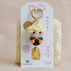 A handmade crocheted keychain featuring a cute character with yellow hair, displayed on a white backing card with Japanese text. The keychain includes a gold-tone ring, a small gold coin charm, and a yellow bag charm, set against a soft beige fabric background.