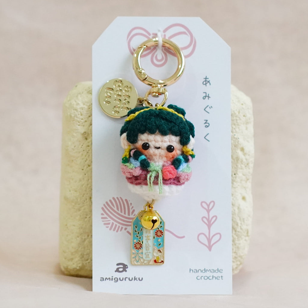 A handmade Japanese-style doll keychain charm with a gold-tone key ring, displayed on a white card backing. The doll features dark green hair, a pink and red outfit, and a small blue and gold hanging charm.