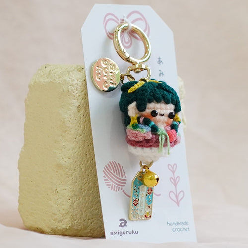 A small enamel keychain with a character charm and gold-tone ring, displayed on a white backing card that stands upright on a textured beige surface.