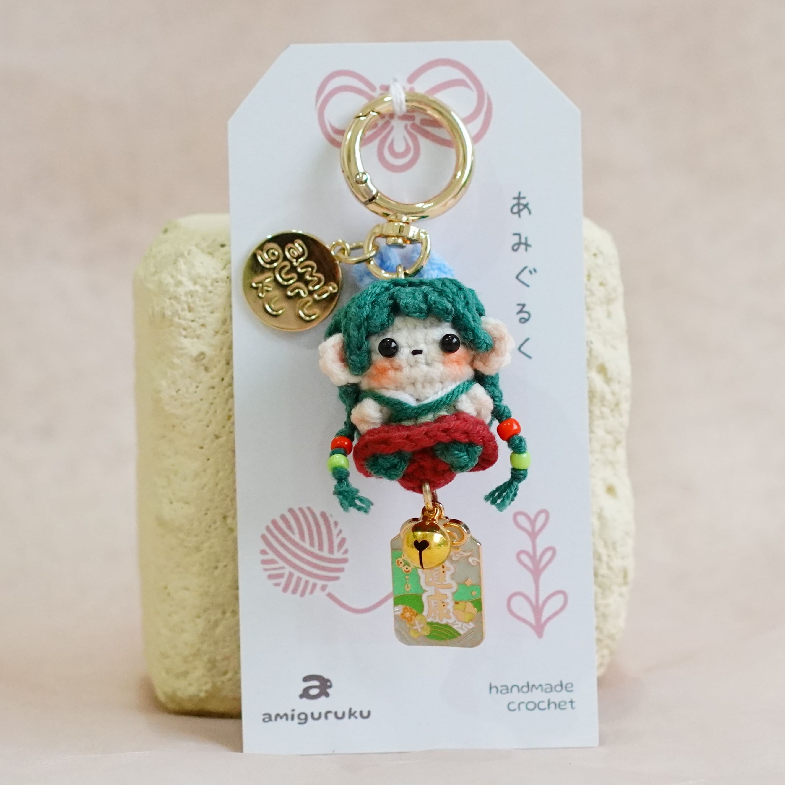 A handmade crochet keychain featuring a small doll with teal hair and a red dress, attached to a white display card with Japanese text and a gold key ring, set against a soft beige fabric background.