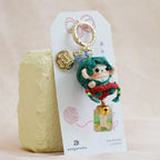 A small handmade doll keychain with green hair, a red outfit, and a gold bell charm, attached to a white paper tag and standing on a pale yellow block against a soft beige fabric backdrop.