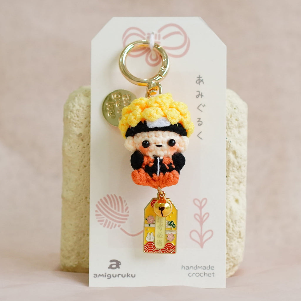 A small Japanese kagura doll keychain with a yellow hat and orange outfit, attached to a white display card and standing in front of two textured cream blocks on a soft beige fabric background.