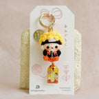 A small Japanese kagura doll keychain with a yellow hat and orange outfit, attached to a white display card and standing in front of two textured cream blocks on a soft beige fabric background.
