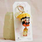 A small crocheted keychain charm with a cartoon face and colorful outfit, attached to a white display card and standing upright on a textured beige surface.