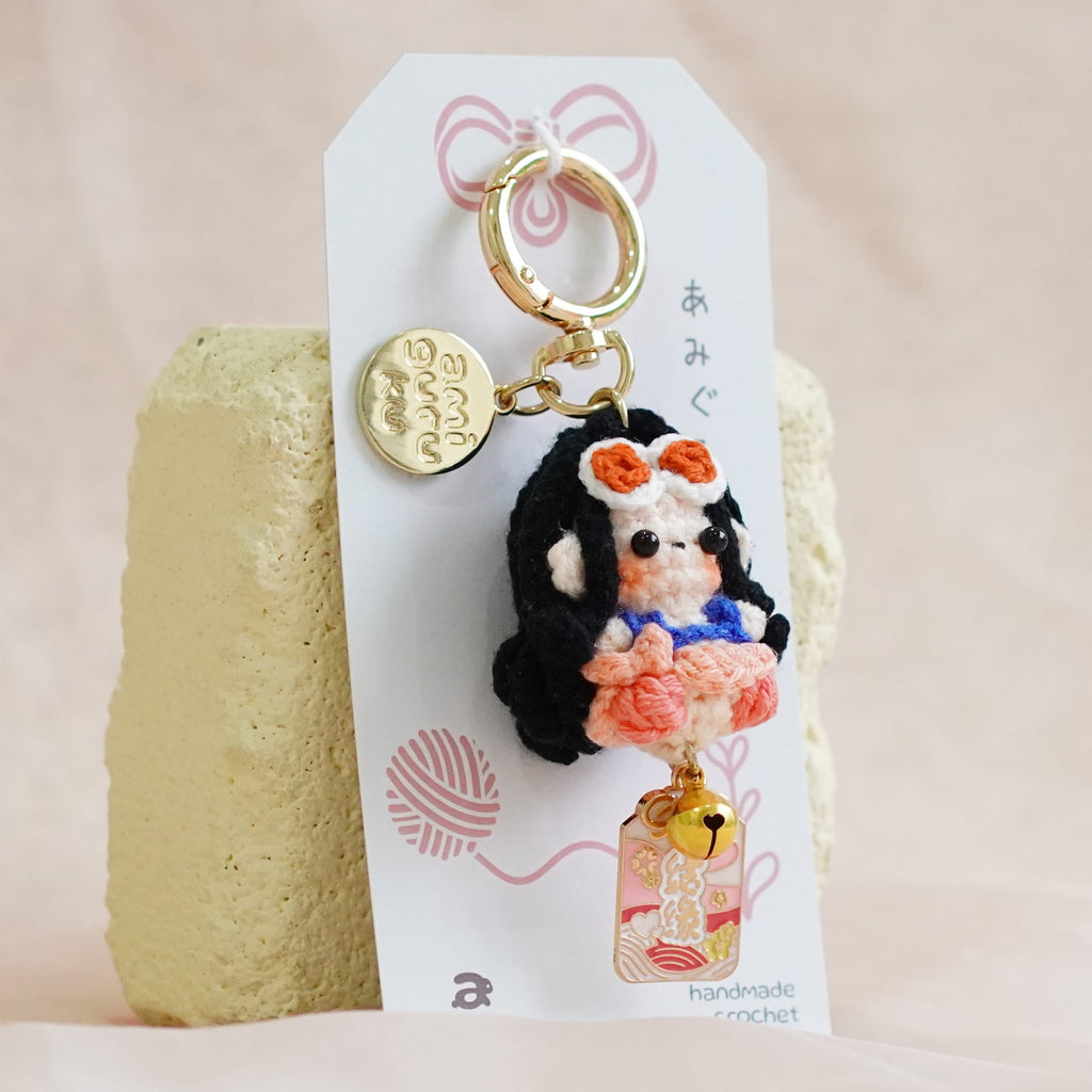 A small handmade doll keychain charm with a gold key ring and a white hanging card, displayed on a textured beige fabric background.