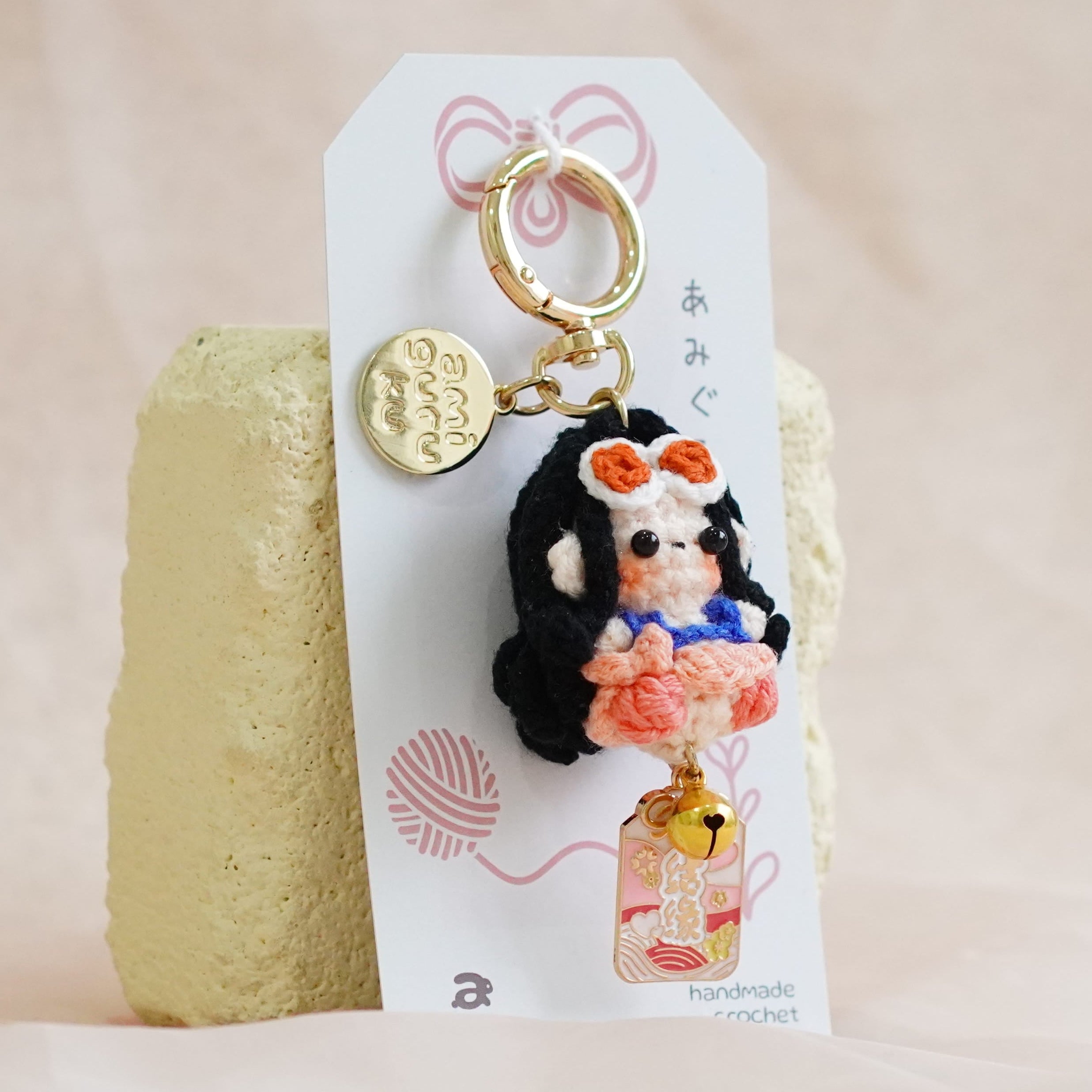 A small handmade doll keychain charm with a gold key ring and a white hanging card, displayed on a textured beige fabric background.