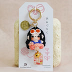 A handmade keychain featuring a small doll charm with black hair, a pink dress, and a gold coin charm, displayed on a white card with Japanese text against a soft beige fabric background.