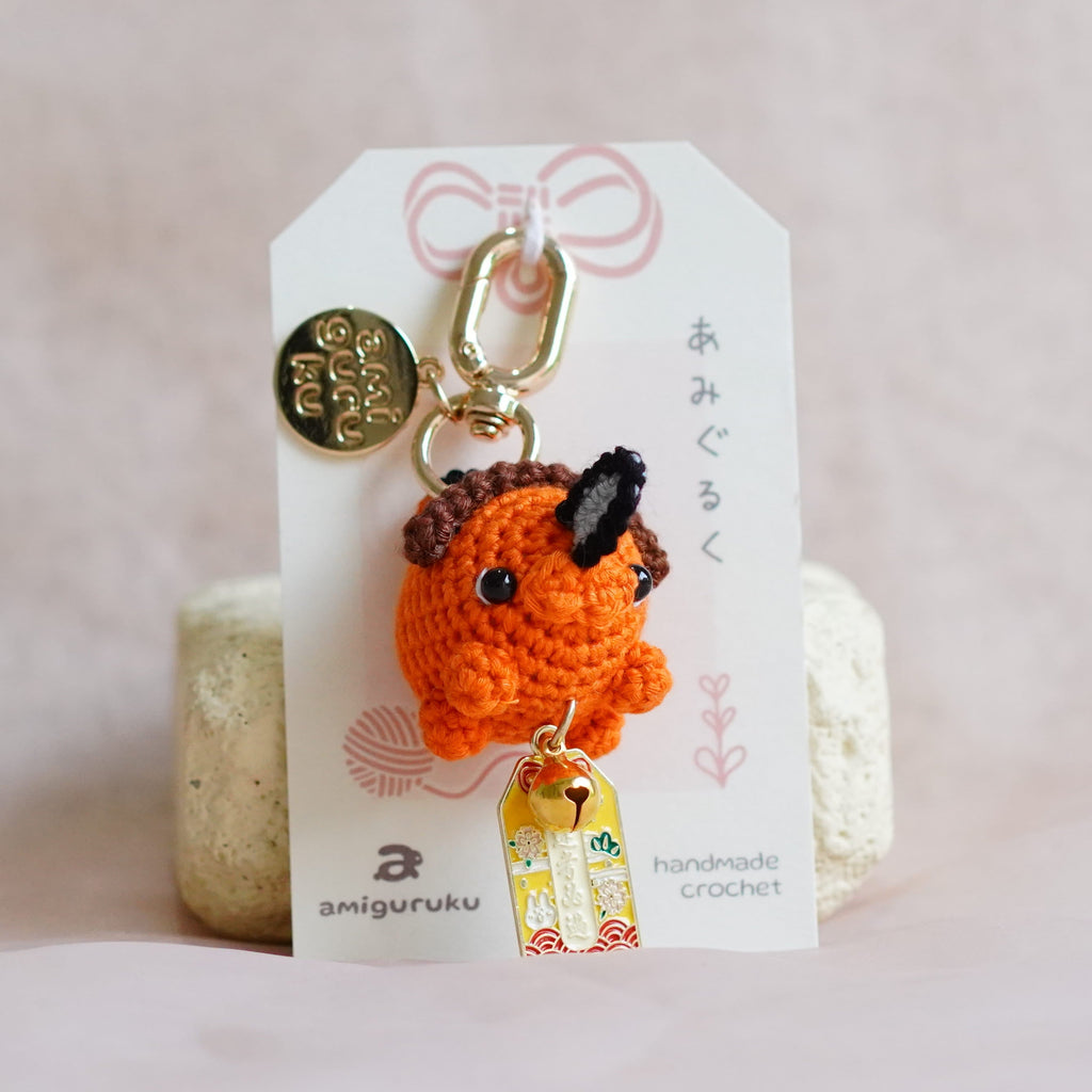 An orange crocheted Pochita keychain with black embroidered eyes and nose, attached to a white card with Japanese text and a small gold coin charm, displayed on a soft pink fabric background.