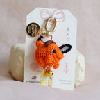 An orange crocheted Pochita keychain with black and white accents, attached to a white display card with a gold clasp, resting on small beige blocks against a soft pink fabric background.