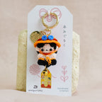 A small anime-themed keychain charm displayed on a white branded card, resting on a textured cream block against a soft beige fabric background.