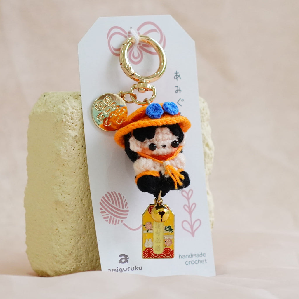 A small character keychain with a gold-tone ring and charm, attached to a white card, standing upright against a soft beige fabric backdrop.