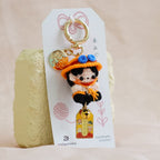A small character keychain with a gold-tone ring and charm, attached to a white card, standing upright against a soft beige fabric backdrop.