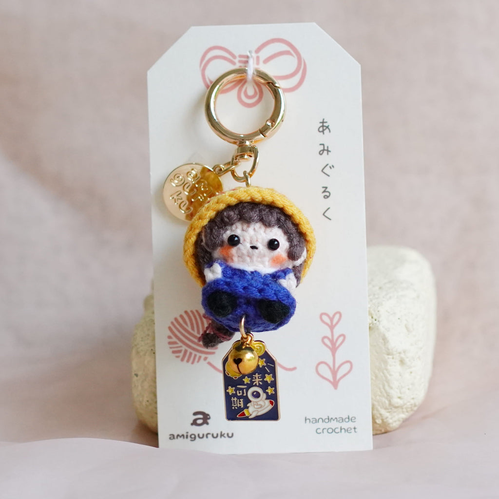 A handmade doll charm keychain displayed on a white card, set against a soft, light pink fabric background.
