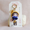 A handmade doll charm keychain displayed on a white card, set against a soft, light pink fabric background.
