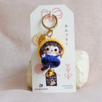 A handmade doll charm keychain displayed on a white card, set against a soft, light pink fabric background.