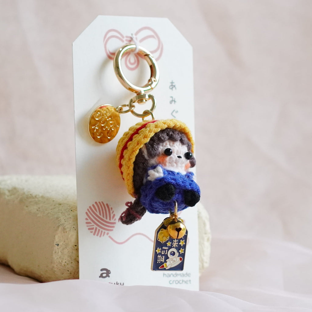 A small plush keychain featuring a cat character wearing a yellow hat and blue outfit, attached to a white display card and standing on a soft beige fabric surface.