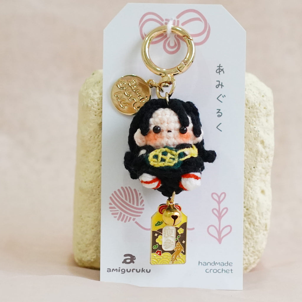 A handmade keychain charm featuring a cute cartoon character with black hair and a yellow top, attached to a white display card with Japanese text and a gold key ring, set against a soft beige fabric background.