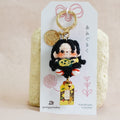 A handmade keychain charm featuring a cute cartoon character with black hair and a yellow top, attached to a white display card with Japanese text and a gold key ring, set against a soft beige fabric background.