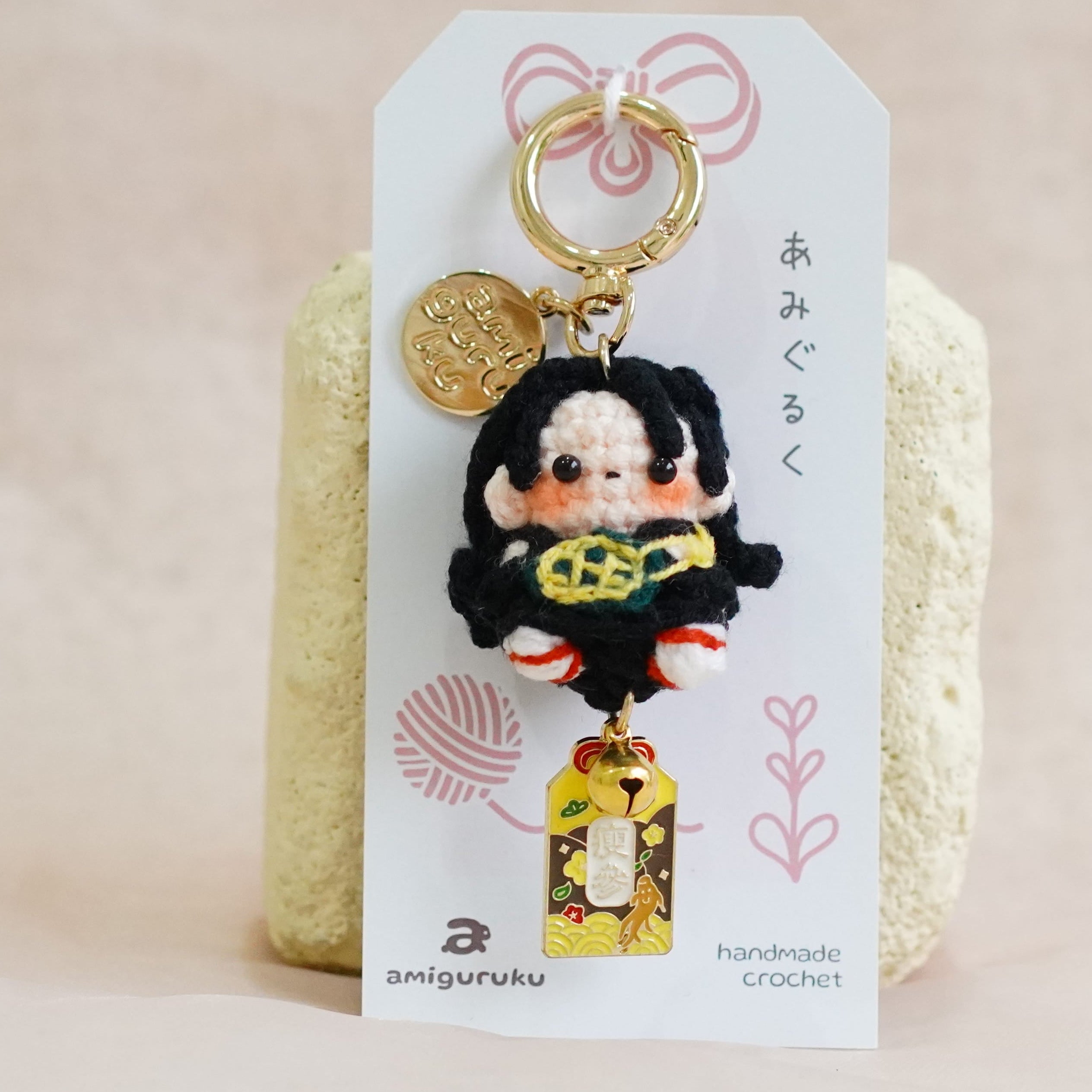 A handmade keychain charm featuring a cute cartoon character with black hair and a yellow top, attached to a white display card with Japanese text and a gold key ring, set against a soft beige fabric background.