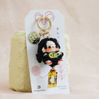 A small plush keychain or bag charm displayed on a white hanging card, resting against a textured pale yellow block on a soft beige fabric surface. The charm features a black-haired character in a black outfit holding a yellow lantern, with a gold key ring and small decorative charms.